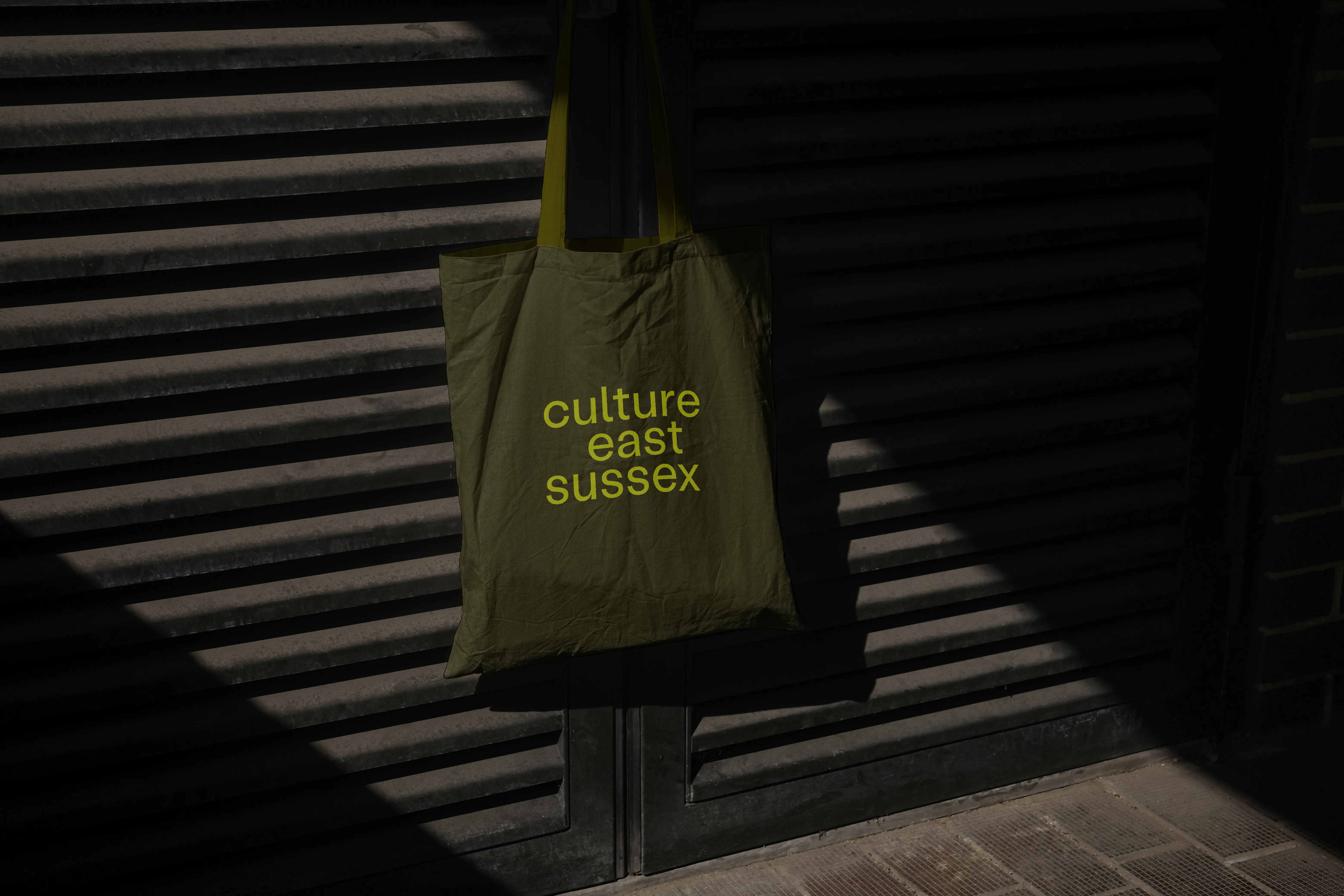 Mock up of a dark olive tote bag showing the Culture East Sussex logotype in acid yellow