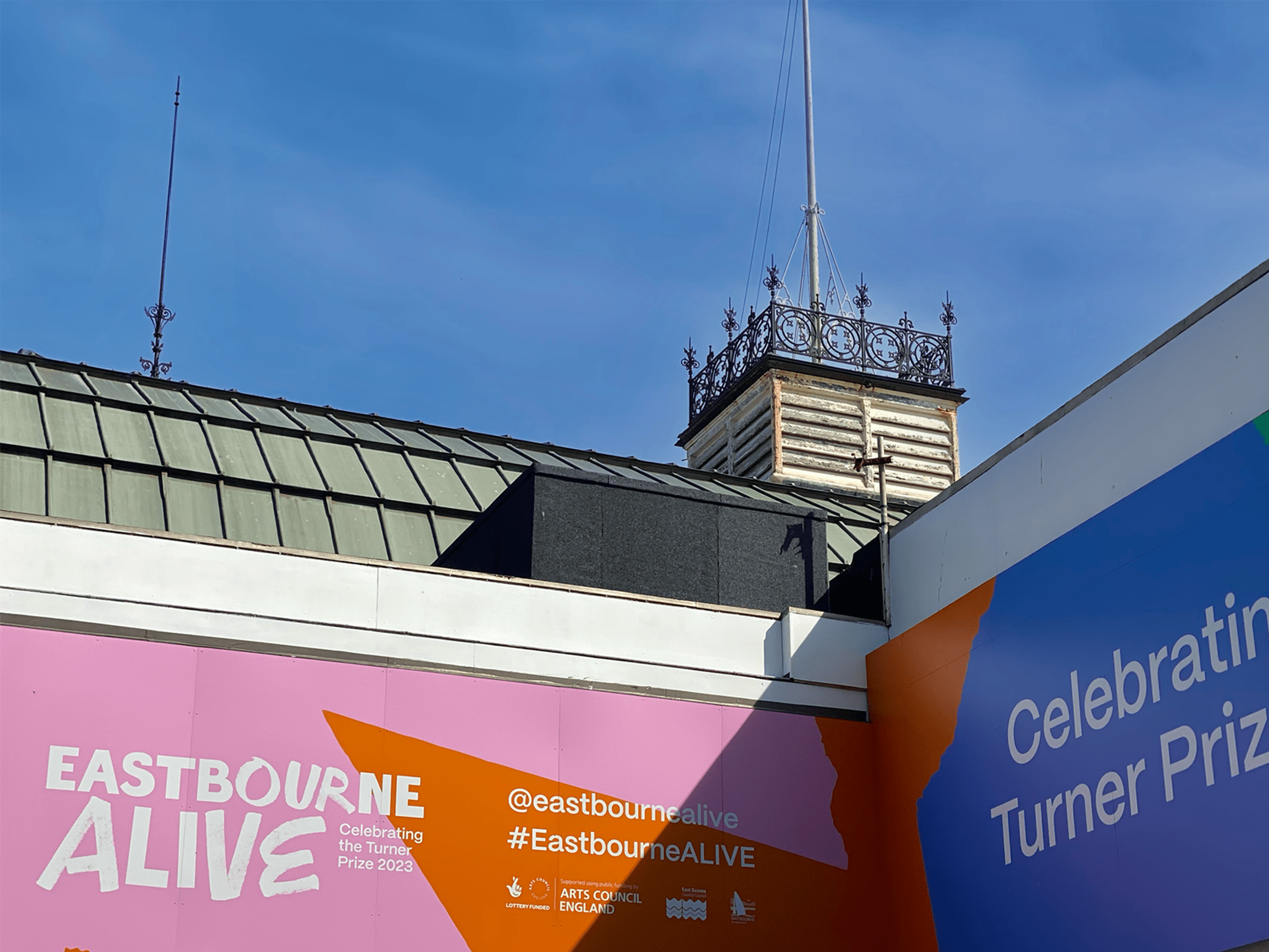 Corner of the Eastbourne Alive hordings at the Eastbourne Winter Garden, showing supper logos in white and the words 'Celebrating the Turner Prize 2023'
